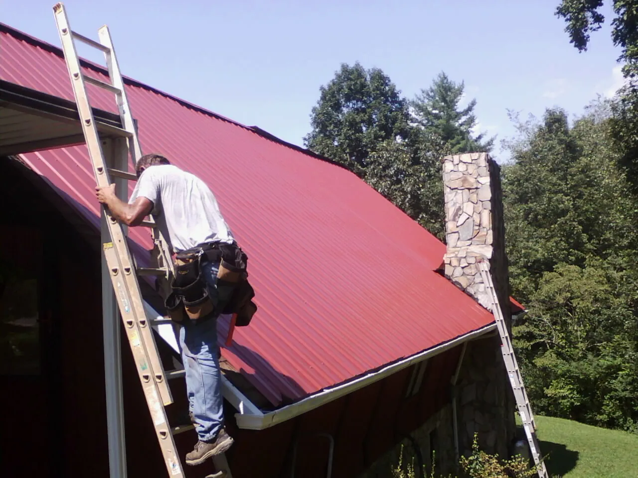 Roofer on a ladder beside a red metal roof and stone chimney — Soffit & Fascia Repair by Roofing Brothers in Shawano